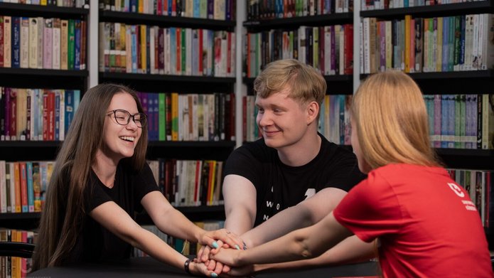 Kolorowe zdjęcie przedstawiające troje uśmiechniętych studentów siedzących na tle bibliotecznego księgozbioru. Na zdjęciu znajdują się dwie kobiety i jeden mężczyzna. Cała trójka trzyma się za ręce w znaku gotowości do wspólnego podjęcia wyzwania. Zdjęcie ma ciepły i jednocześnie motywacyjny charakter